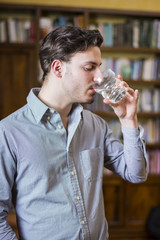 Young man in shirt drinking water from crystal glass on background of bookshelves. 