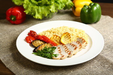 Grilled chicken with slices, with vegetables, bulgur on a white plate, decorated with parsley. In the background, lettuce leaves, color pepper. Low-calorie, dietary food for a healthy lifestyle