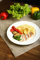 Grilled chicken with slices, with vegetables, bulgur on a white plate. In the background leaves and pepper. Low-calorie, dietary food for a healthy lifestyle. Wooden table, burlap, dark background