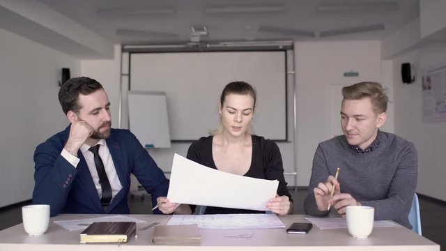 At the table young group of three works on a drafting project. Female business leader distributes a mock-up on paper for designer and architect to create an engineering solution for building for