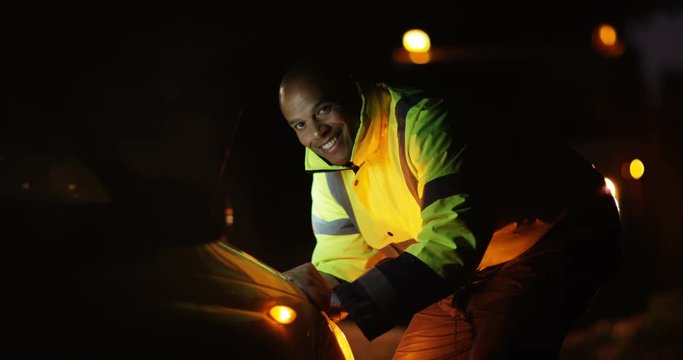 4K Portrait smiling mechanic looking at engine of broken down car at night. Slow motion.