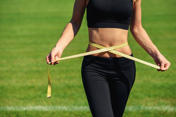 Young fitness girl in black sportswear with perfect waist with a measuring tape in hands against...