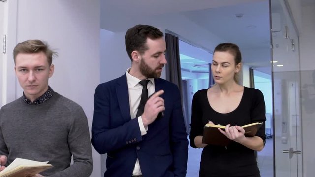 Businessman is giving tasks to his male and female interns in the office. Three people are going in a row through the office corridor and boss in blue suit and tie is talking to woman in black dress