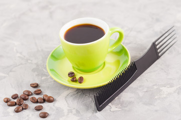Single new plastic comb near full cup of black tasty coffee and lot of scattered fried coffee beans on green round ceramic saucer on old gray broken concrete