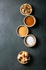 Salted caramel fudge candy served with fleur de sel, caramel sauce, brown cane sugar and caramelized walnuts in wood bowls over black texture background. Top view, space. Dessert set