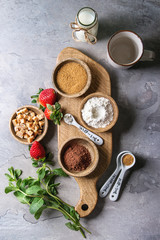 Ingredients for cooking chocolate mug cakes. Flour, cocoa powder, sugar, caramel in wooden bowls, milk, strawberries and mint served with spoons and mug over grey texture background. Top view, space