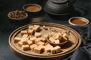 Salted caramel fudge candy served on wooden board with caramel sauce, brown cane sugar and caramelized walnuts in wood bowls, iron teapot over black texture background. Close up.