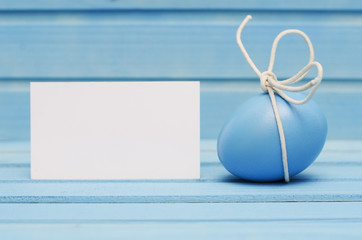 Blue Easter egg with white bow on blue wooden background with blank card