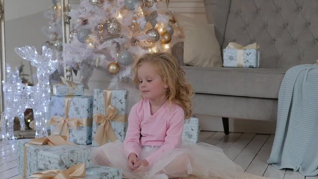 the girl sits on the floor of the house with Christmas gifts and a garland. Girl playing with Christmas gift