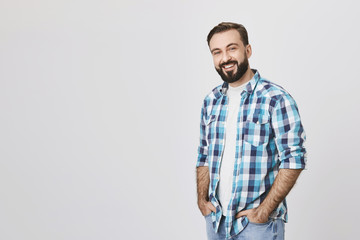 Adult bearded pleasant guy standing half turned with hands in jeans, smiling happily against gray...