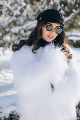 Fototapeta na wymiar A beautiful, stylish, fashionable woman in a fur coat, hat and glasses, posing in the park in snowy weather.