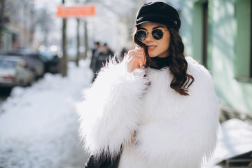 Fototapeta na wymiar A beautiful, stylish, fashionable woman in a fur coat, hat and glasses, posing on the street in snowy weather.