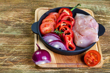 Cooking chicken. Raw chicken breast fillet with red sweet pepper, red onion, tomatoes and rosemary, in the black pan, prepared for cooking. Selective focus, horizontal.
