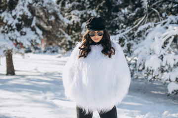 Fototapeta na wymiar A beautiful, stylish, fashionable woman in a fur coat, hat and glasses, posing in the park in snowy weather.