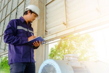 Asian engineer or technician in blue collar safety uniform and white security helmet checking motor gear machinery equipment and writing data on notebook with sun light effect for industrial concept