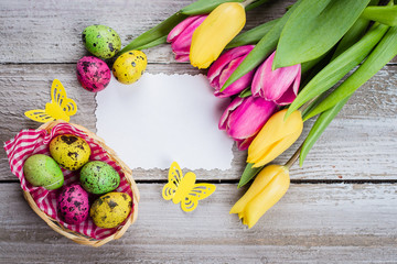 Spring Greeting card with empty paper blank. Easter Background with yellow and pink tulips and colorful quail eggs in nest on wooden table background. Top view, flat lay, copy space.
