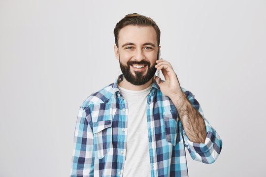 Studio Shot Of Attractive Cheerful European Guy With Beard And Moustache, Talking Via Smartphone And Smiling Broadly To Camera, Standing Over Gray Background. Man Is Happy He Changed Mobile Operator