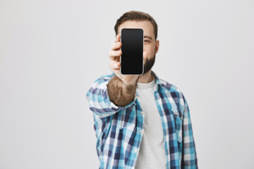 Male model with beard and trendy haircut in checked shirt showing smartphone at camera covering face with it, standing against gray background. Consultant shows gadget to customer of shop he works at