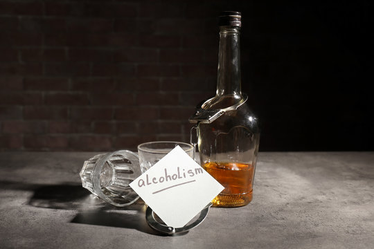 Note with word "Alcoholism", bottle of alcohol and handcuffs on table