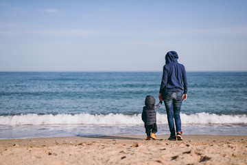 Young woman standing at sea sand beach background holding hand little cute child baby boy, looking on horizon. Mother, little kid son. Family day 15 of may, love, parents, children concept. Back view.