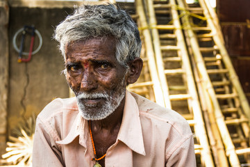 Portrait of Indian elder man with traditional bindi as a third eye, white beard, bamboo ladders on the background and a saw hanging from the wall in Mysore, Karnataka, India. Rural village hard worker