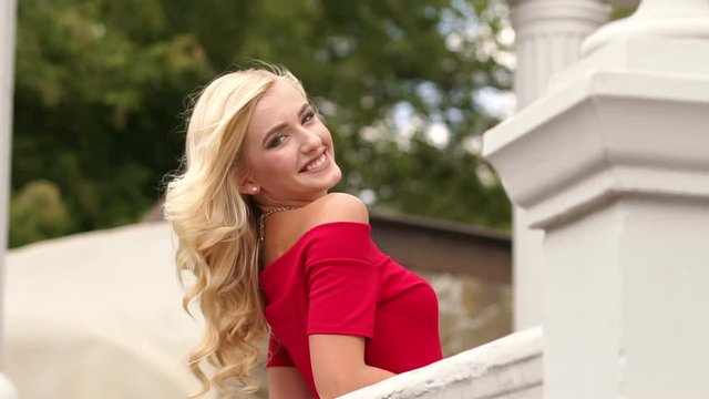 Beautiful young woman in red dress with long flowing hair stands leaning on the railing of the old vintage ladder. Slow motion. Portrait of beautiful girl with long flowing hair.