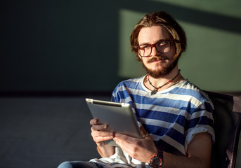 Smiling teenager in glasses using tablet, standing in a dark room near the window, indoor shot in the sunlight