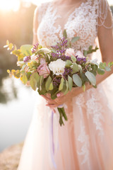 The bride in a beige wedding dress holding a lush bridal bouquet of lilac roses and a lot of greenery. Stylish wedding bouquet on the sunset background
