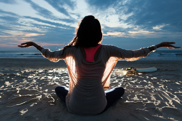 Indian surfer girl with long hair sitting on the beach in lotus pose embracing last sunset light...