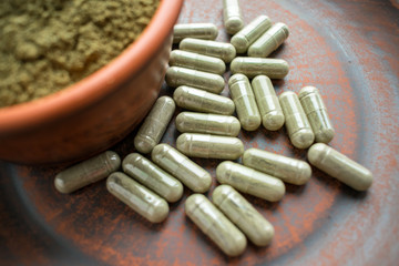 Supplement kratom green capsules and powder on brown plate. Herbal product alt-medicine kratom is  opioid. Home alternative pain remedy, opioid addiction, painkiller, overdose. Selective focus