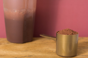 Whey Protein. Front view of scoop with chocolate powder and shaker on rustic wooden table. Solid color: brown.
