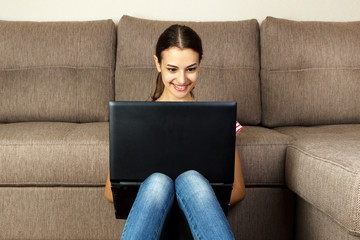Naklejka premium Beautiful girl in casual clothes is using a laptop and smiling while sitting on the floor near the couch at home