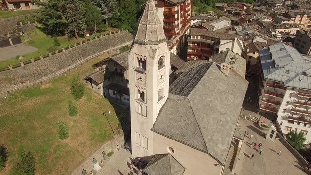 4k Old ancient church bell tower tolling for Sunday mass people waiting to go into. Aerial from above top view of small Italian town Courmayeur tiles roofs Aosta Valley at foot of Mont Blanc mountain