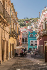 A charming cobblestone pedestrian street with colorful buildings, outdoor table, and a hill dotted with colorful homes, in the background, in Guanajuato, Mexico