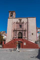 Front shot of an old stone church, with a bell tower and a red double sided staircase, inside the Plaza San Roque, with paving stones and a clear, deep blue sky, in Guanajuato, Mexico - 191672563