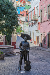 The statue in the Plaza del Ropero, with decorative stone pavement, partial view of a tree, and a narrow street heading up to colorful houses dotting the hillside, in Guanajuato, Mexico - 191673378