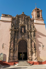 The front exterior of El Templo de San Diego, with elaborately decorative stone entryway, bell tower, two light sconces, stone steps, a shadow and deep blue sky, in Guanajuato, Mexico - 191673785