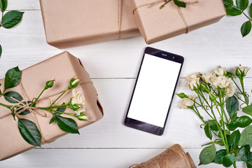 Closeup of mobile smartphone with blank screen, gift boxes and fresh roses on wooden background, copy space. Greeting card mockup. Top view, flat lay. Spring, Mothers day, Womens Day, Easter, birthday
