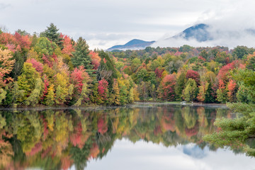 Vibrant fall colored forest with dark mountain behind it.  Both mountain and trees full of fall foliage reflecting in a smooth lake in New England.