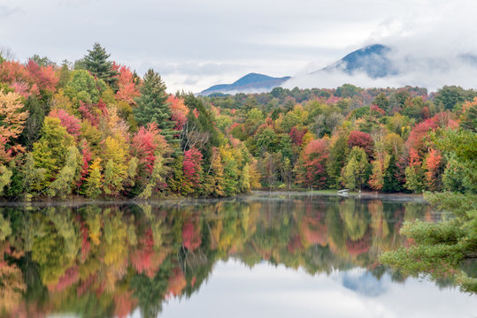Vibrant fall colored forest with dark mountain behind it.  Both mountain and trees full of fall foliage reflecting in a smooth lake in New England.