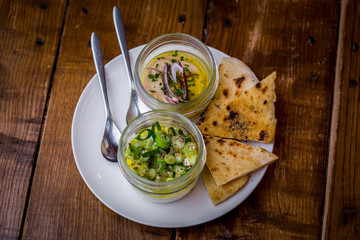 top view of hummus and tahini in glass jar topped with chives onion and herbs with spoon on wooden table 