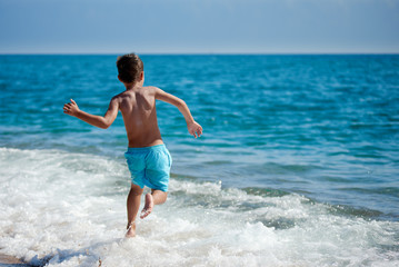 Caucasian boy is running in the water along the sea shore. Back view.