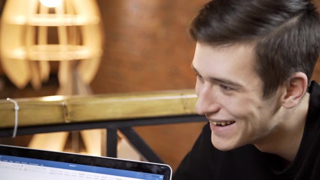 Young man with laptop has good time in cafe indoors. Handsome guy is talking while sitting at table in cozy place. Brunet speaks positively with friend, smiling amiably and laughing, touching face
