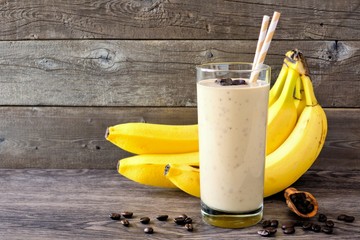 Coffee, banana smoothie in a tall glass with coffee beans and bananas in background. Side view, against a rustic wood background.
