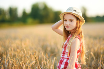 Adorable girl wearing straw hat walking happily in wheat field on warm and sunny summer evening.