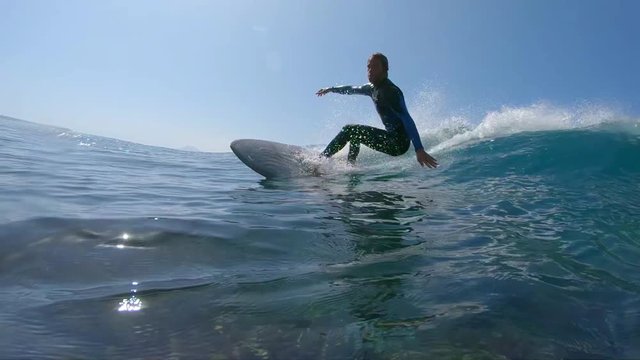 SLOW MOTION, UNDERWATER: Young male surfer carving awesome waves in exotic surf spot in summer sunshine. Fit surfboarder rides and carves approaching breaking wave on his surfboard in sunny seaside.