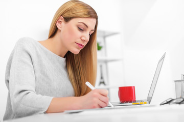 A woman in a white office is sitting at a table and writing. A young lady using a laptop fills documents in the workplace.