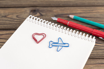 paper clip in the form of a heart, an airplane on a notepad, pen, pencil on a wooden background. romantic trip. plans for recreation.