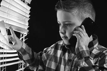 The boy looks out the window through the blinds and talking on the phone