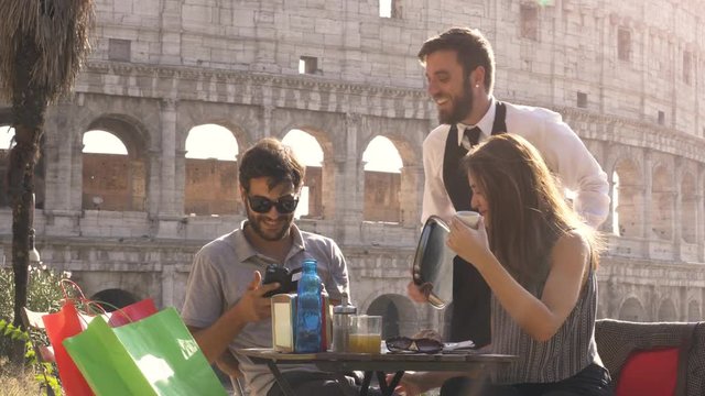 Happy young couple tourists taking selfies with nice italian elegant waiter sitting at bar restaurant in front of colosseum in rome at sunset joking and laughing having fun